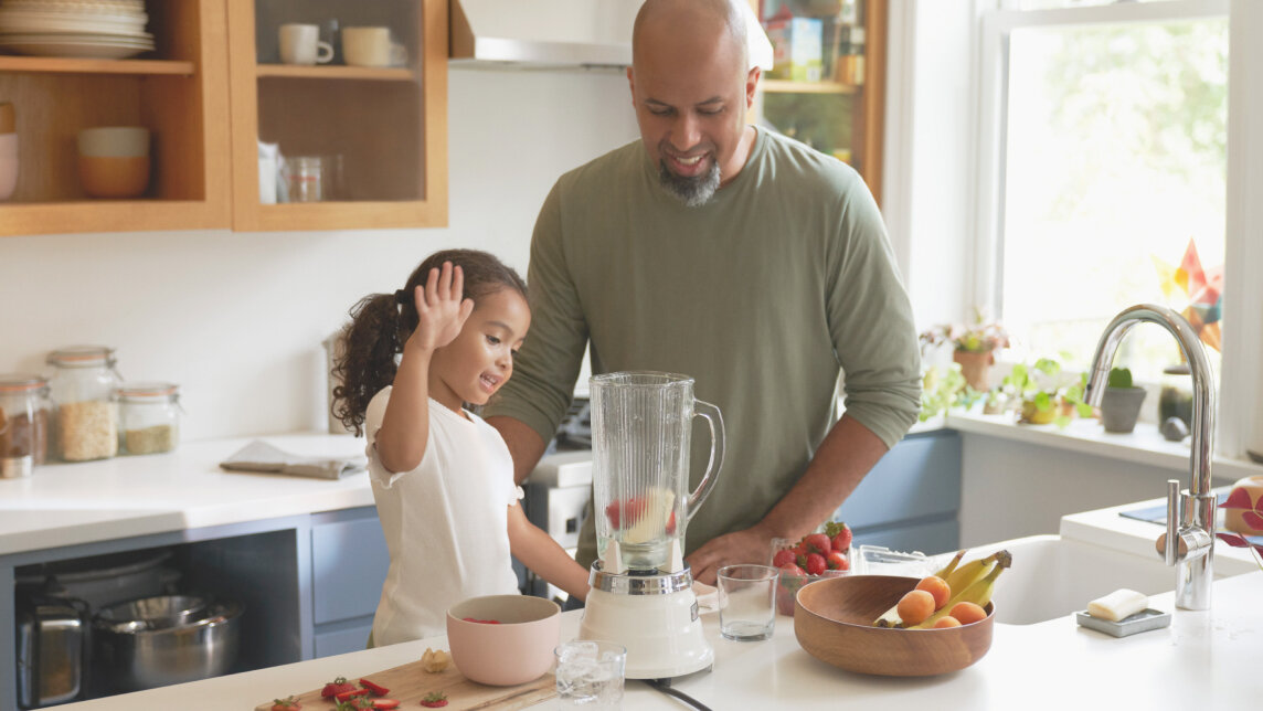 A father and his young daughter in the kitchen on a work island. They are cutting up strawberries and other fruit to prepare a smoothie in an electric blender.