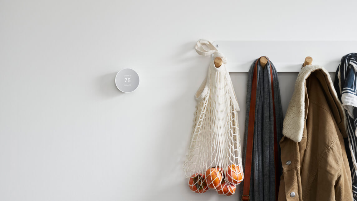 A Nest Thermostat on a white wall, next to a coat rack. On the coat rack, there are winter jackets, scarves and a net filled with mandarins/ oranges.