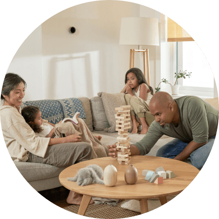 Una mamá, un papá y sus dos hijas pequeñas están jugando en el sofá. El papá está jugando Jenga y la mamá y las niñas lo están mirando.